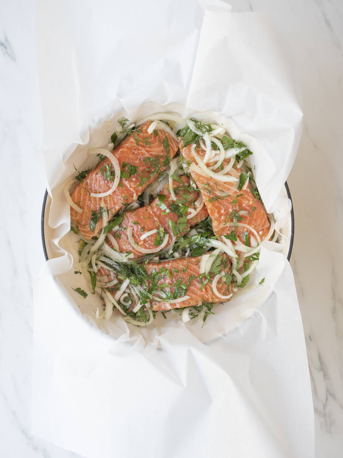 A skillet lined with parchment paper with the salmon fillets topped with herbs and sliced onions.