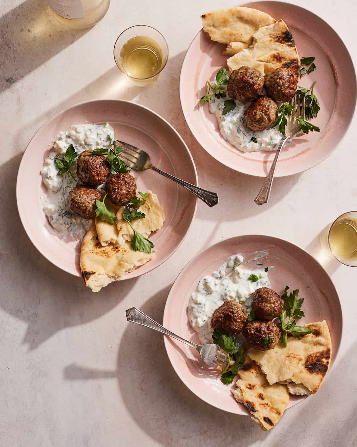 Greek lamb meatballs served in 3 separate pink bowls topped with parsley leaves with a side of naan bread served with two glasses of white wine 