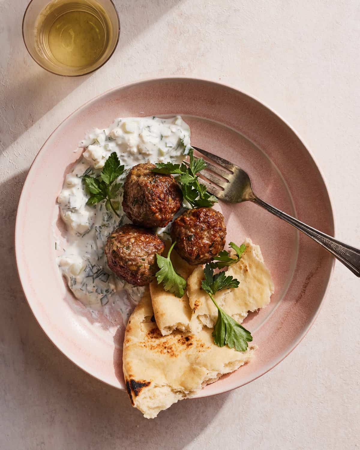 Greek lamb meatballs and tzatziki topped with fresh parsley and served with a side of naan in a pink ceramic bowl with a glass of white wine in the top left corner