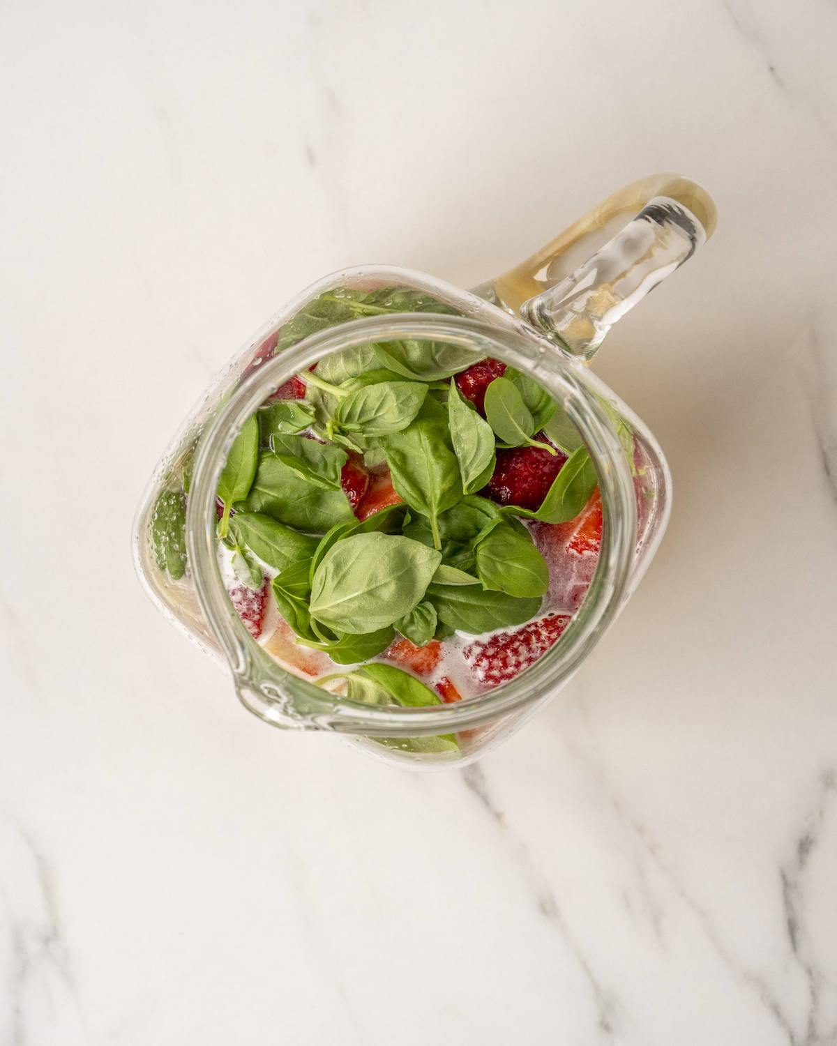 Lemon juice and dissolved sugar with strawberries and basil leaves in a glass pitcher on a white countertop.