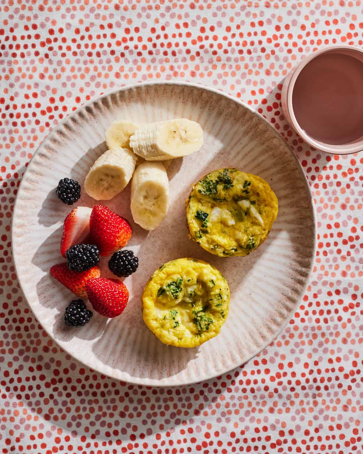 Two zucchini feta frittatas with a side of berries and sliced bananas on a ceramic plate and a cup of water in the top right corner
