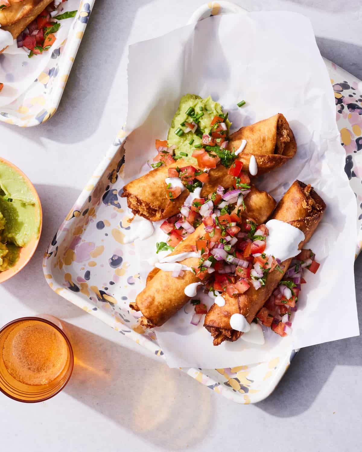 A rectangular plate with parchment paper with three chicken flautas, topped with pico de gallo, sour cream and guacamole, a similar plate on the corner of the image, a small bowl of guac and a glass of water peeping in from the side.