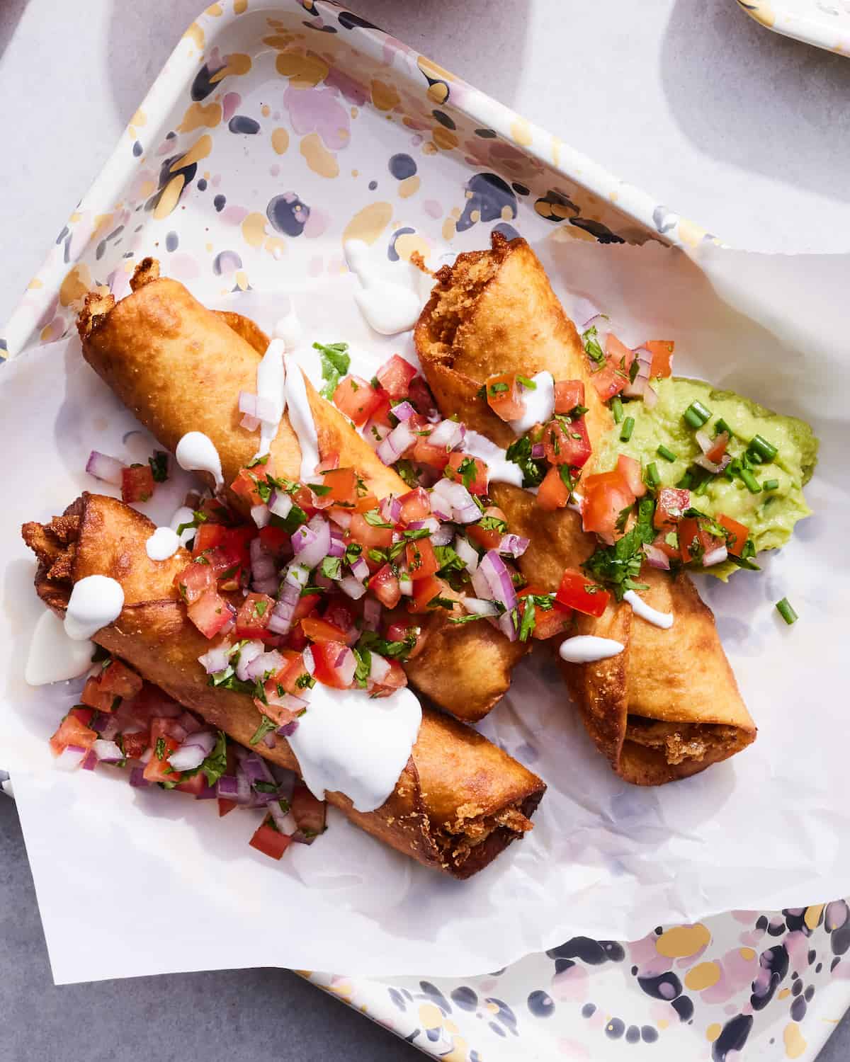 A rectangular plate with parchment paper with three chicken flautas, topped with pico de gallo, sour cream and guacamole.