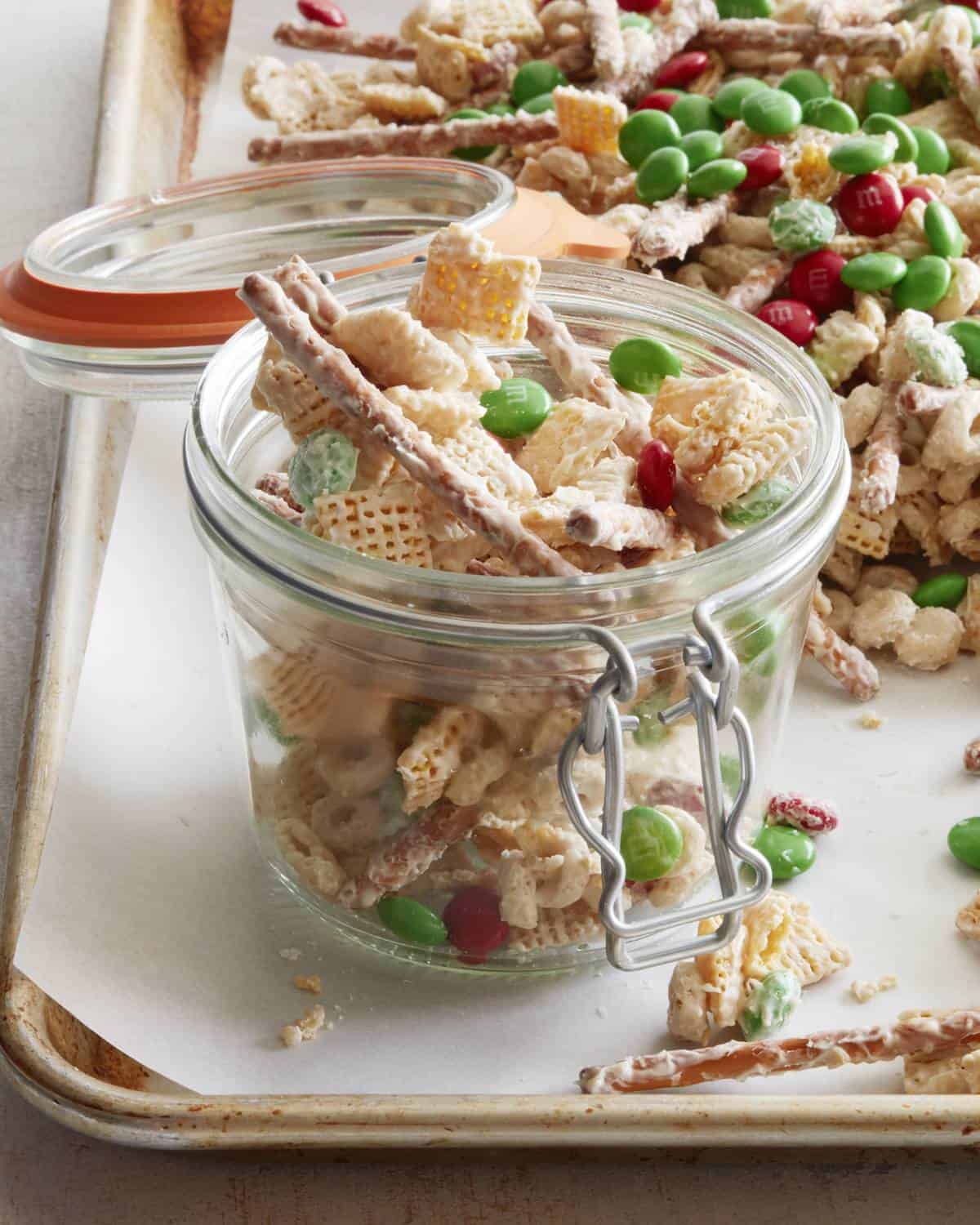 A close-up shot of an open glass canister with christmas chex mix, placed on a parchment-lined baking sheet with more christmas chex mix on it.