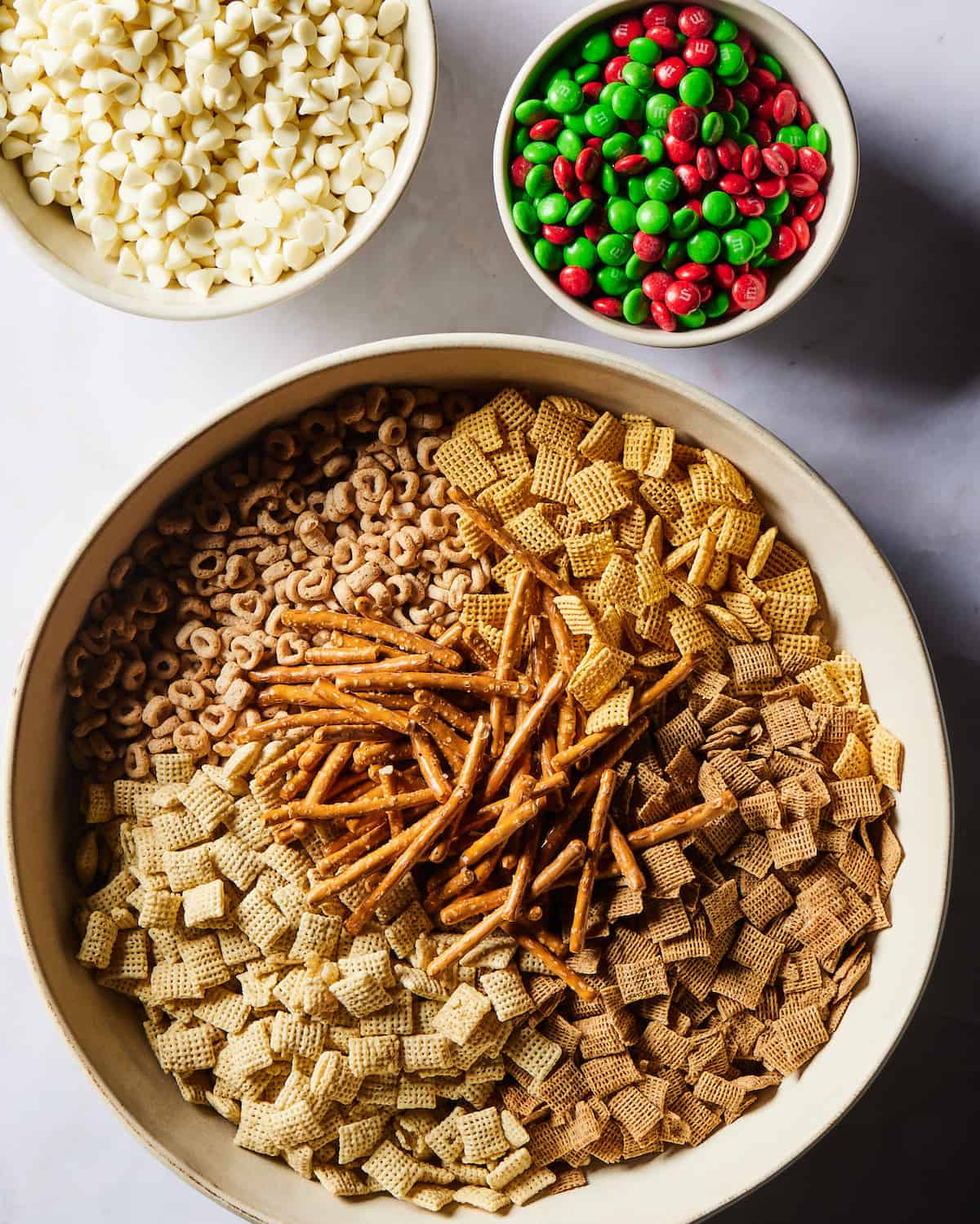 A large white round bowl with corn chex, wheat chex, rice chex, cheerios and pretzel sticks in it, with a small bowl full of red and green M&Ms and another small bowl with white chocolate chips on its side.