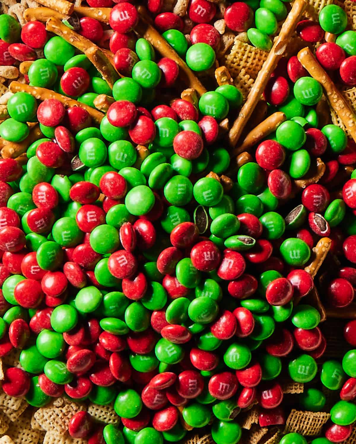 A close-up shot of mixed-up chex, cheerios, pretzel sticks and red and green M&Ms.