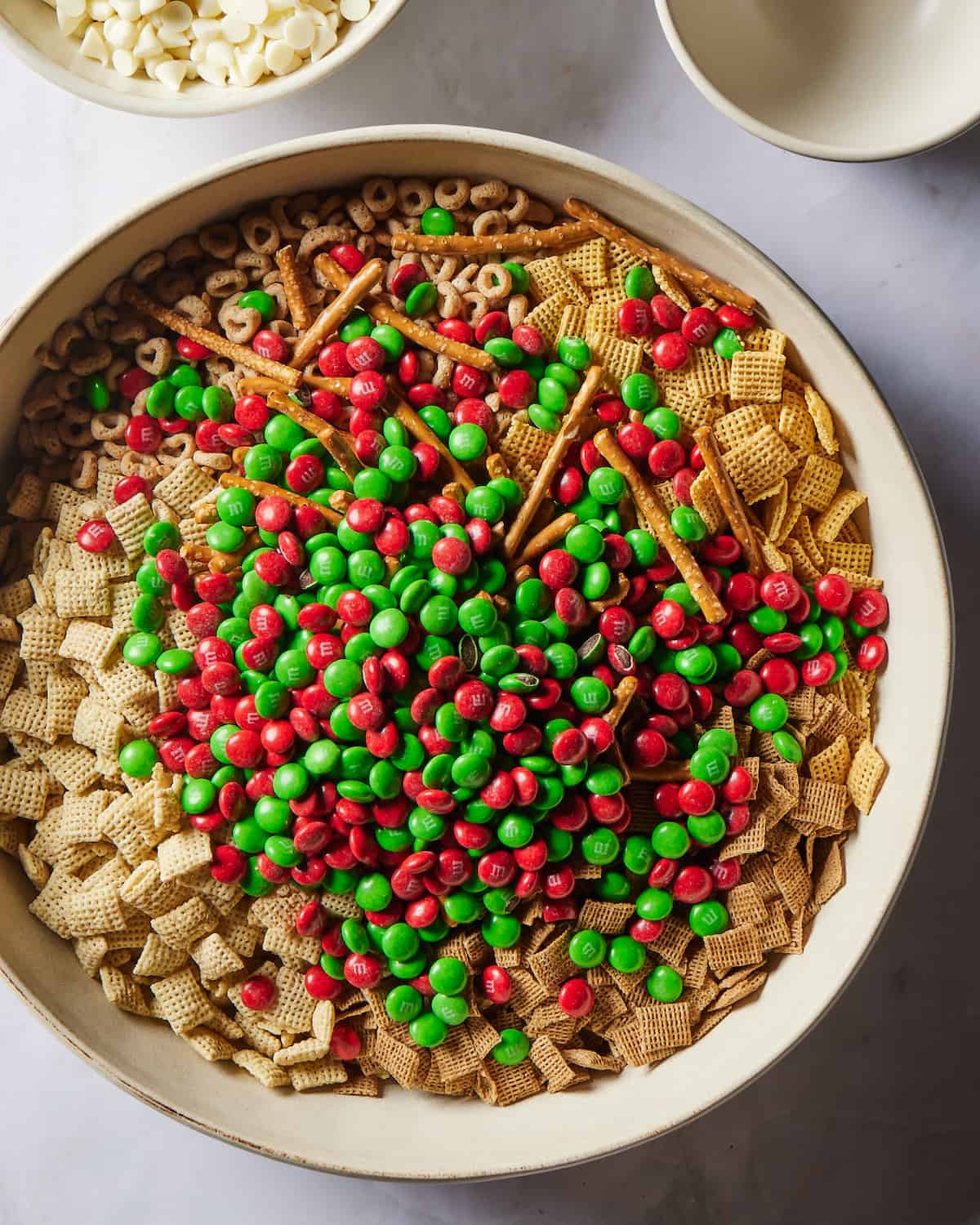 A large white round bowl with corn chex, wheat chex, rice chex, cheerios and pretzel sticks plus red and green M&Ms in it, a small empty bowl and another small bowl with white chocolate chips on its side.
