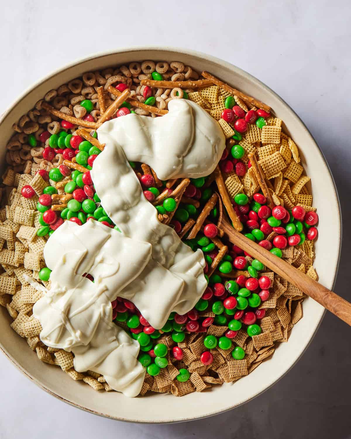 A large white round bowl with corn chex, wheat chex, rice chex, cheerios and pretzel sticks plus red and green M&Ms in it, topped with molten white chocolate drizzled on top and a wooden spoon.