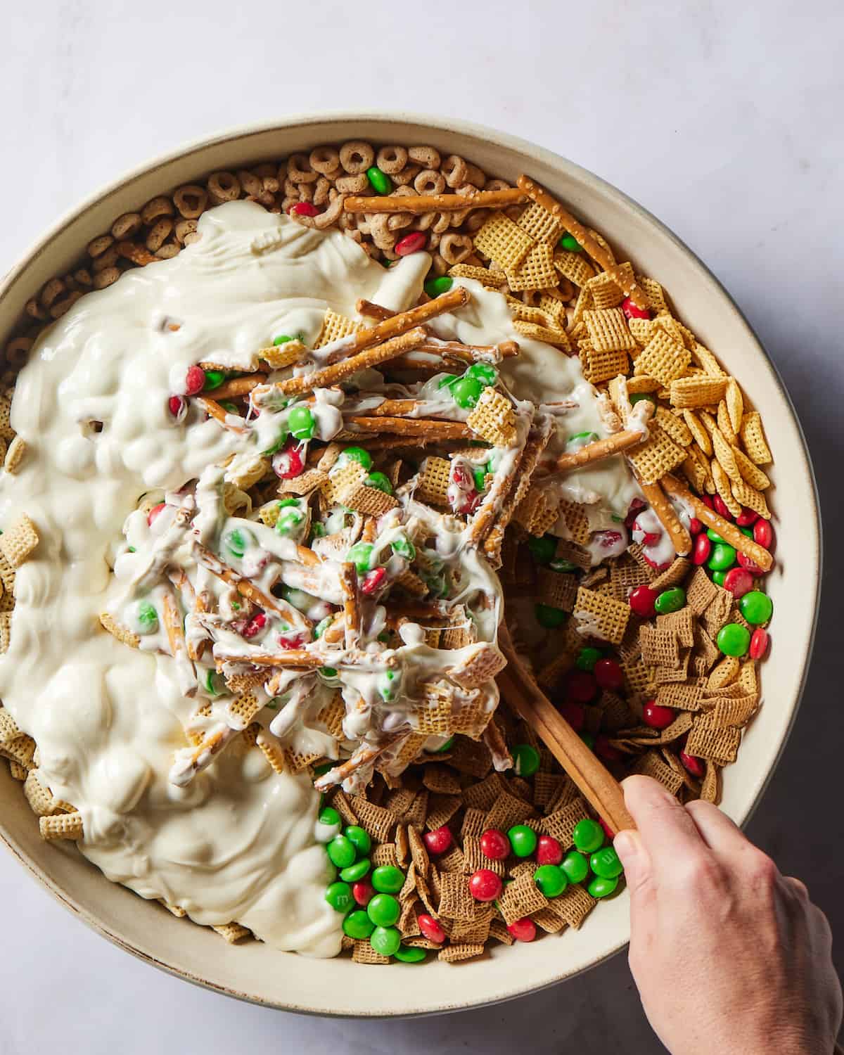 A large white round bowl with corn chex, wheat chex, rice chex, cheerios and pretzel sticks plus red and green M&Ms in it, topped with molten white chocolate drizzled on top and a woman's hand using a wooden spoon to mix the white chocolate in.