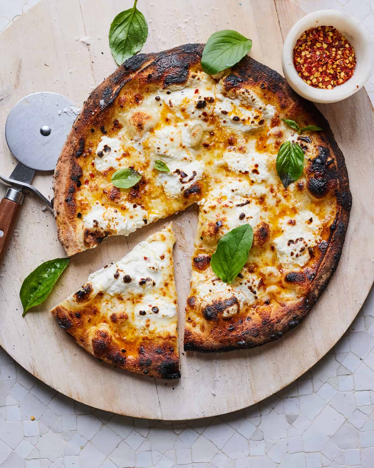 Cooked Pizza Bianca topped with basil leaves on a wooden pizza peel with a pizza cutter to the left of the pizza and red pepper flakes in a white marble bowl