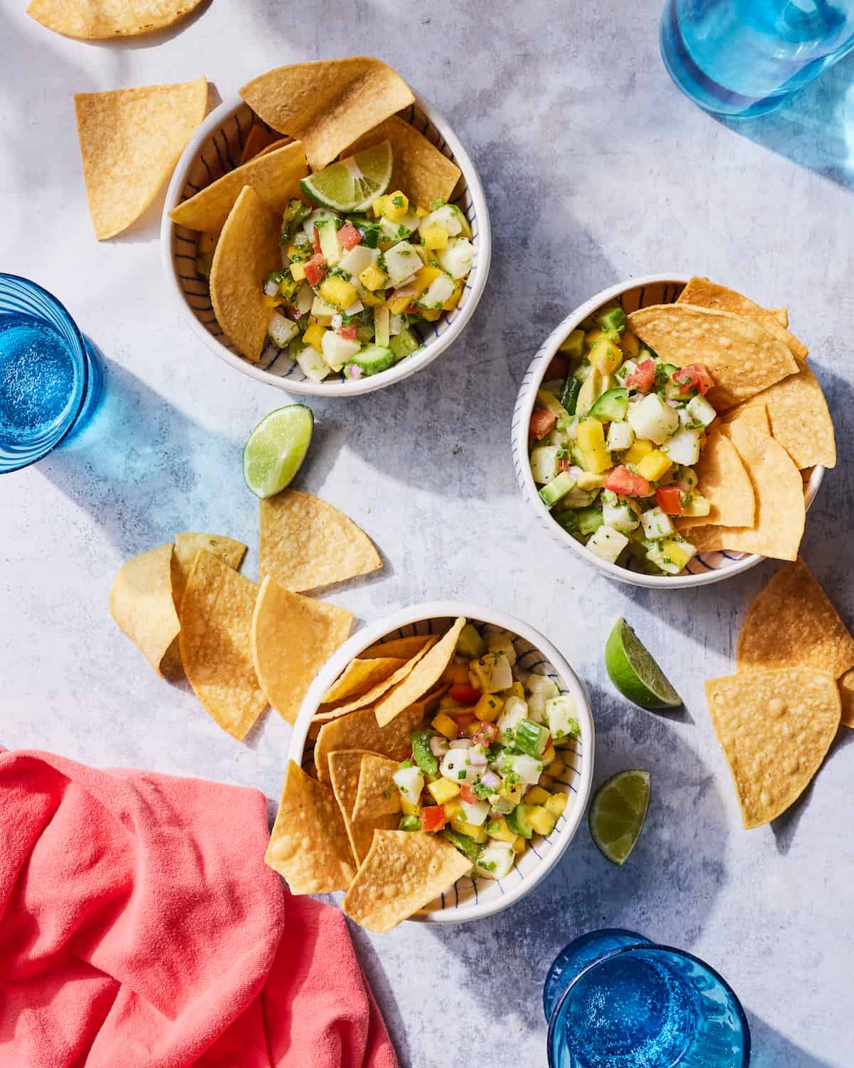 Three ceramic bowls filled with Sea Bass Ceviche and tortilla chips beside tortilla chips, lime wedges, a pink linen napkin in lower left corner, and three blue glasses of water in upper right and left corner and the lower right corner on a countertop.