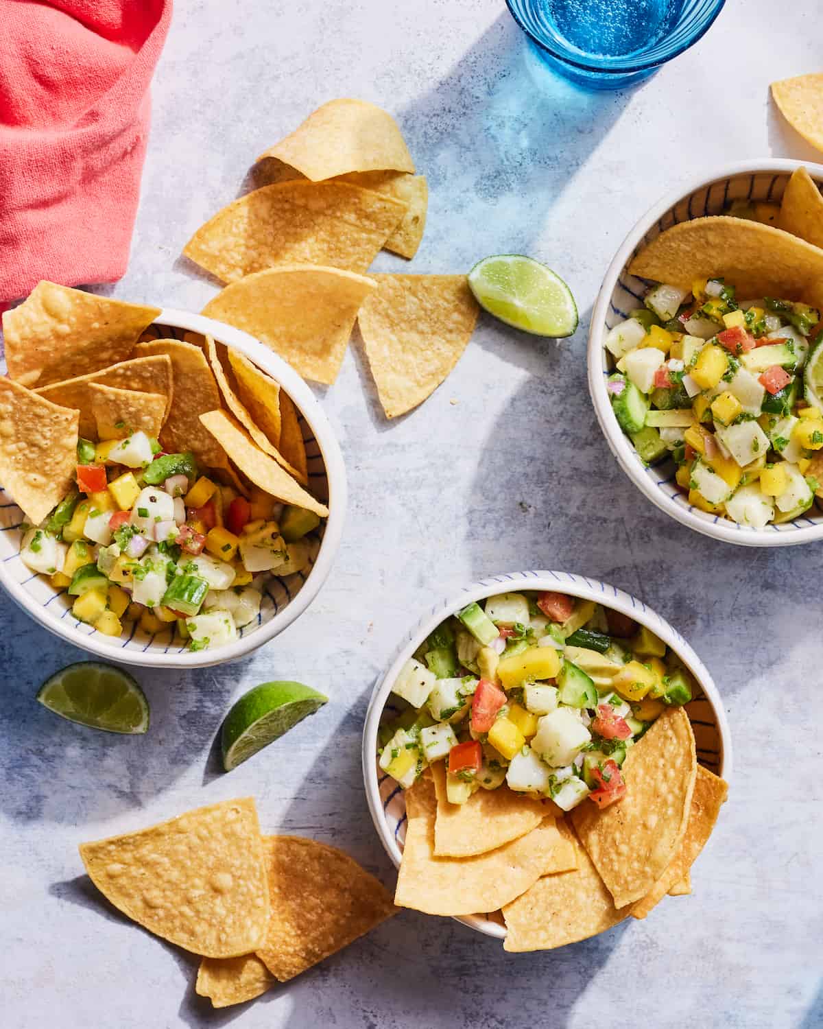 Three ceramic bowls filled with Sea Bass Ceviche and tortilla chips beside tortilla chips, lime wedges, a pink linen napkin in upper left corner, and a blue glass of water in upper right corner on a countertop.