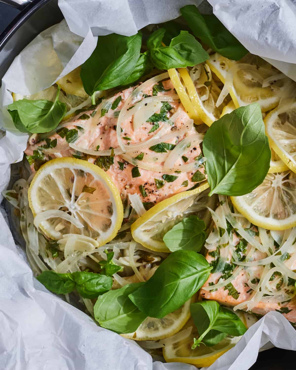 A close-up shot of a skillet lined with parchment paper, with a salmon placed inside, topped with thinly sliced lemon slices, onion slices and lots of herbs and basil leaves.