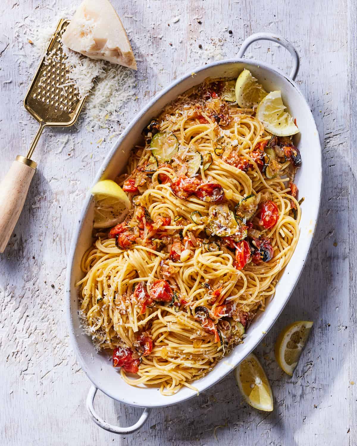 A serving dish of Summer Ratatouille Pasta topped with lemon wedges beside lemon wedges and a cheese grater with grated parmesan cheese in the top left corner.