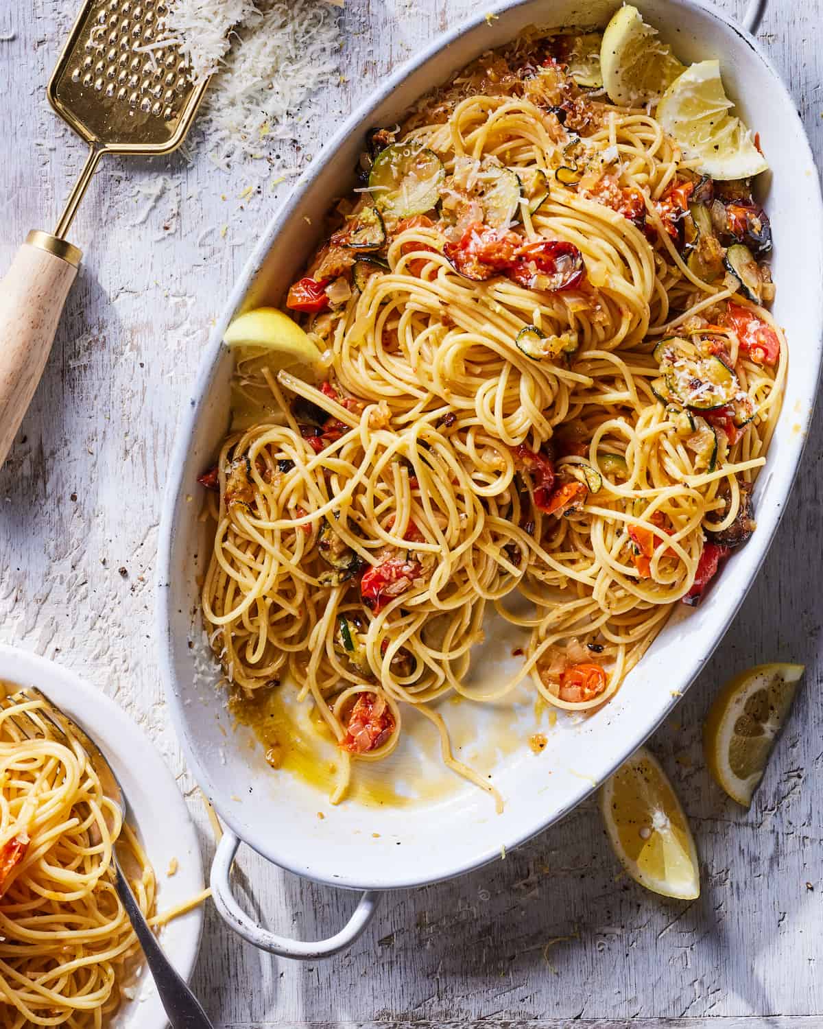 A serving dish of Summer Ratatouille Pasta topped with lemon wedges beside a bowl of a serving of the Summer Ratatouille Pasta and lemon wedges and a cheese grater with grated parmesan cheese in the top left corner.