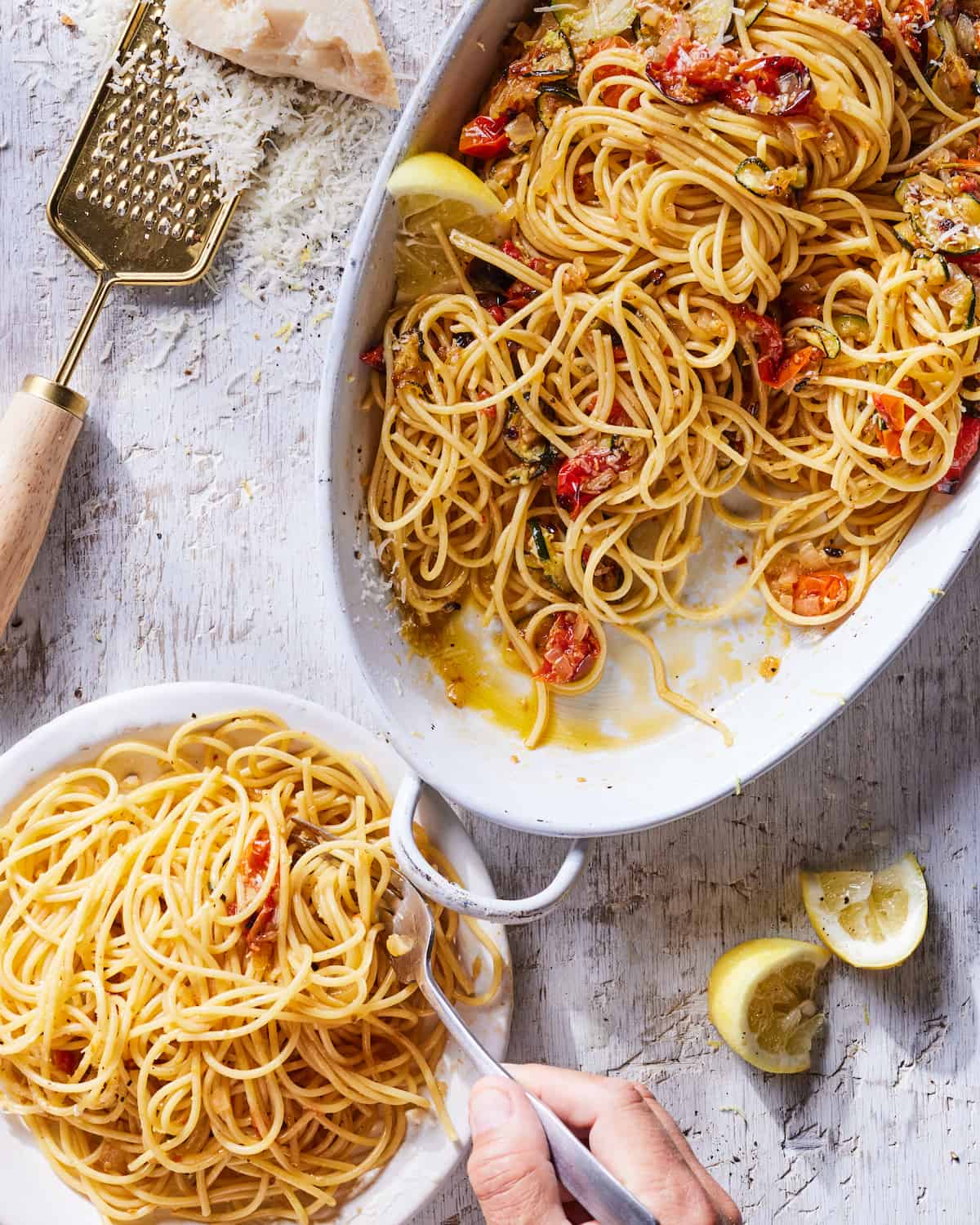A serving dish of Summer Ratatouille Pasta topped with lemon wedges beside a bowl of a serving of the Summer Ratatouille Pasta and lemon wedges and a cheese grater with grated parmesan cheese in the top left corner.