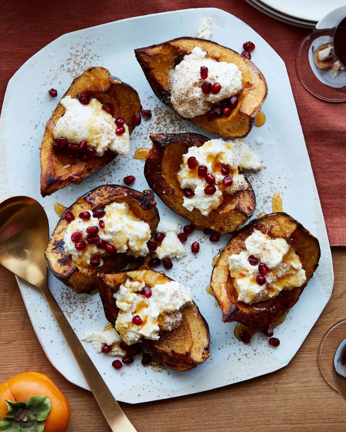A white serving platter with roasted acorn squash topped with ricotta, pomegranate seeds, nutmeg and honey, with a golden serving spoon.