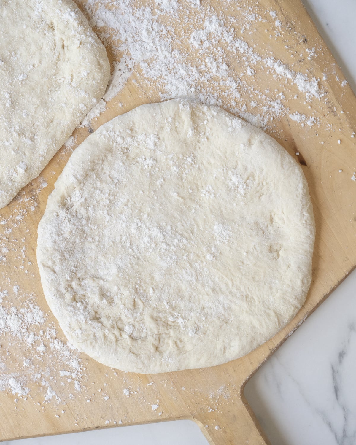 Rolled and shaped pizza dough divided into two pieces on a wooden pizza peel sprinkled with flour