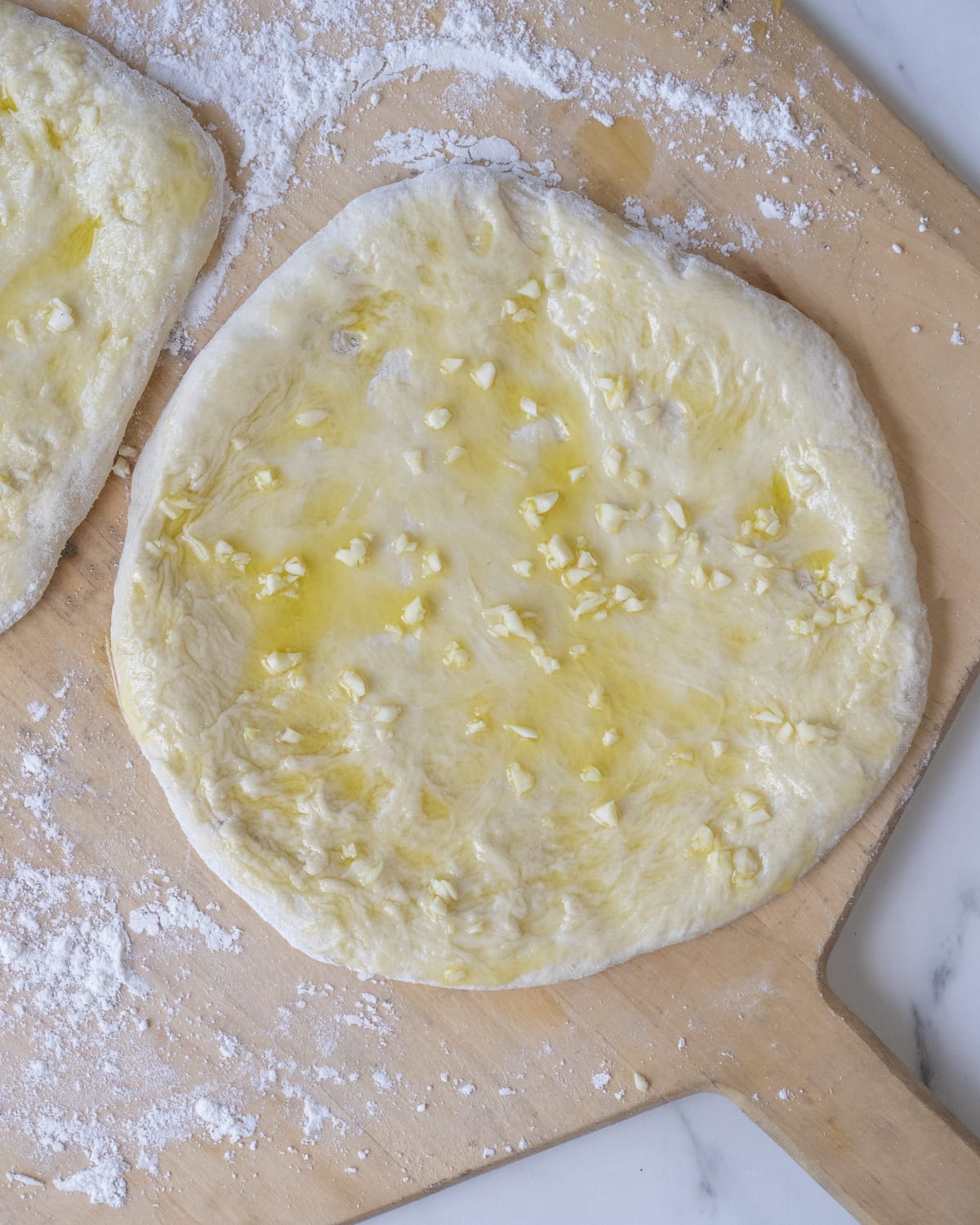 Rolled and shaped pizza dough divided into two pieces topped with olive oil and finely chopped garlic on a wooden pizza peel sprinkled with flour