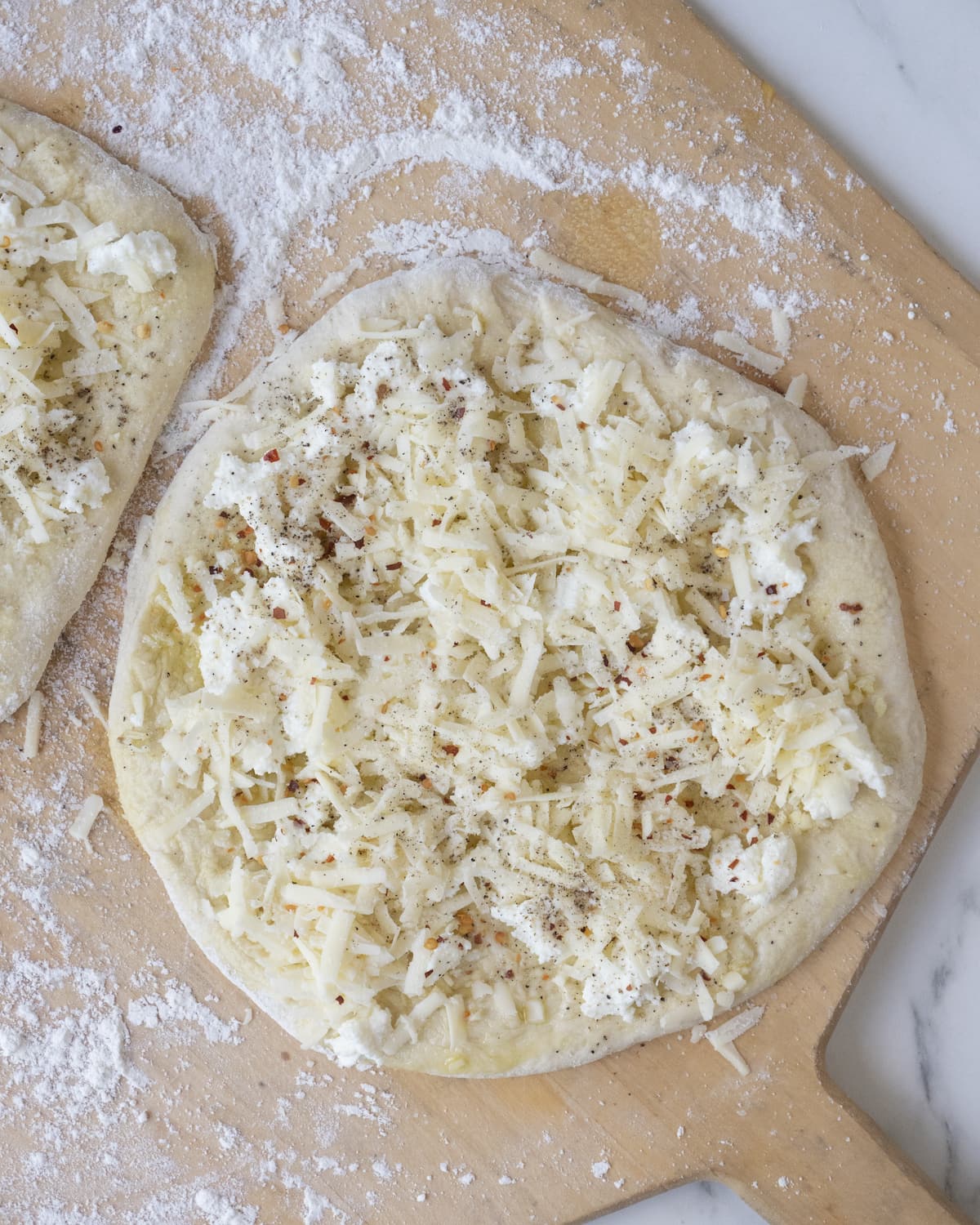 Rolled and shaped pizza dough divided into two pieces topped with olive oil and finely chopped garlic and cheeses and red pepper flakes on a wooden pizza peel sprinkled with flour
