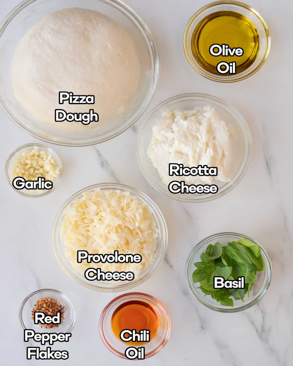 Ingredient shot of ingredients in individual clear bowls including red pepper flakes, basil leaves, chili oil, provolone, and ricotta cheese, garlic, pizza dough, and olive oil