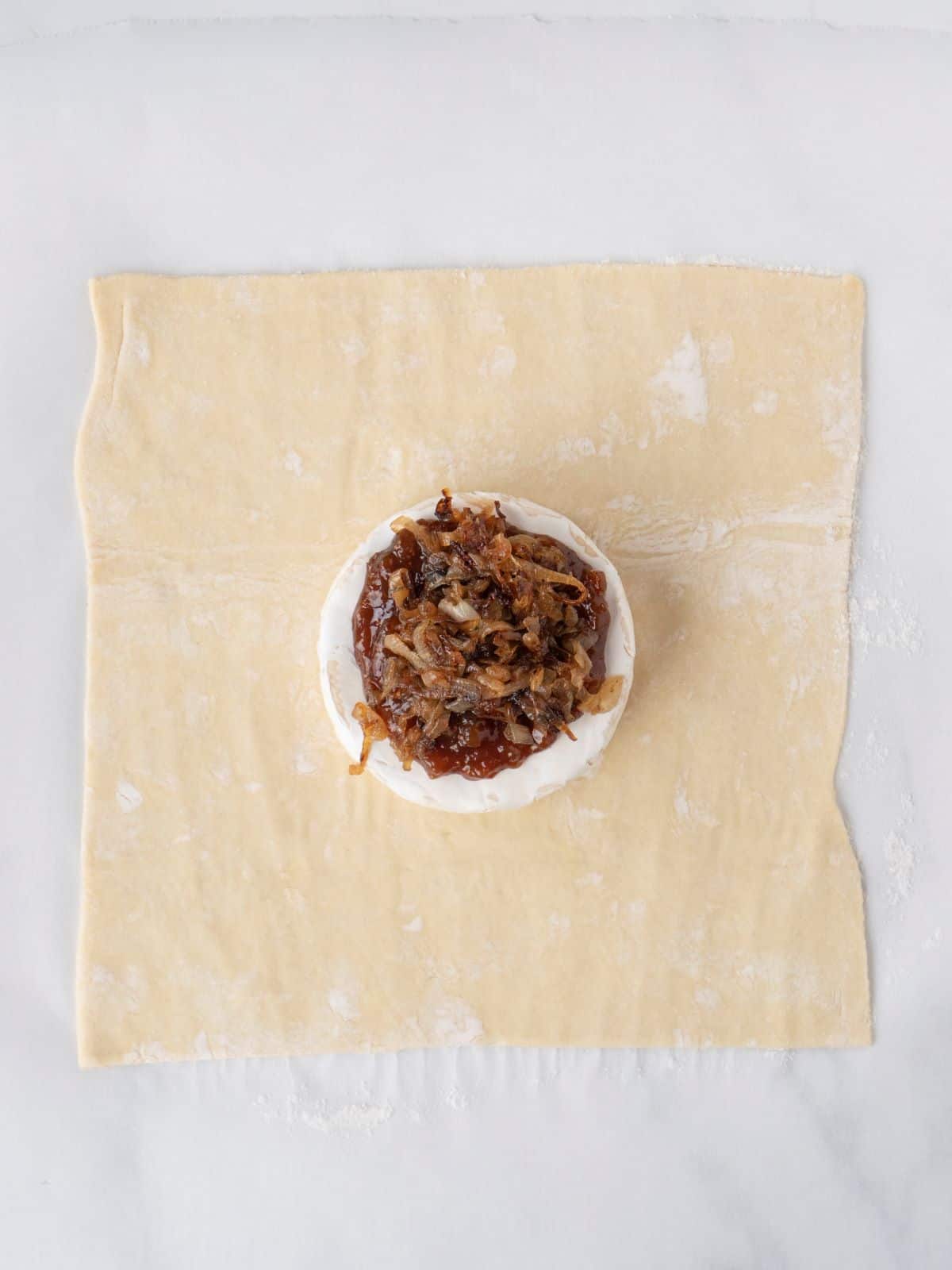 A rolled out puff pastry with a wheel of brie topped with caramelized onion and fig jam in the center.