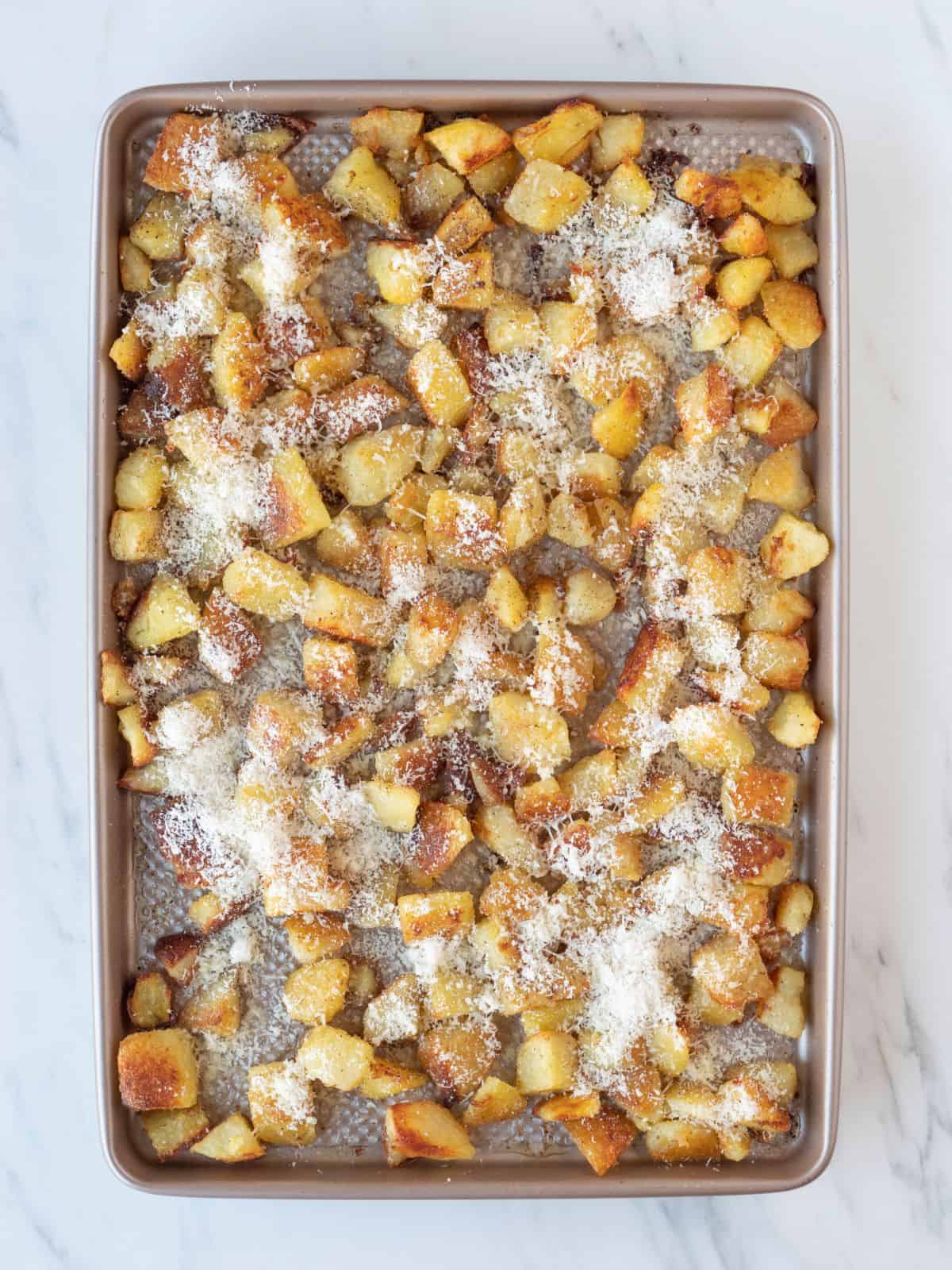 A baking sheet with boiled cubed potatoes on it that have been roasted in the oven and are golden with cheese sprinkled on top.
