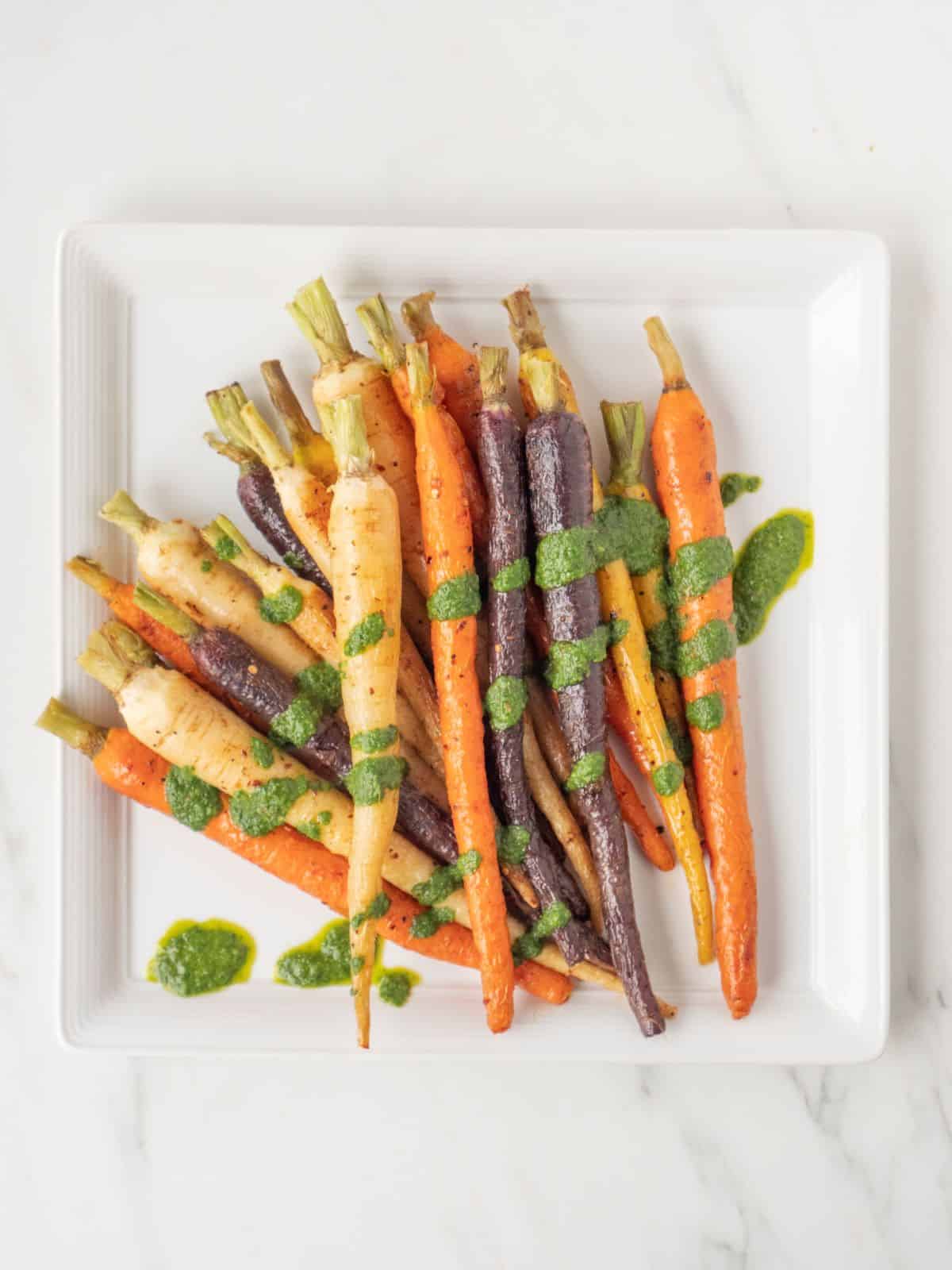A square white platter with charred rainbow carrots and basil mint sauce drizzled on top.