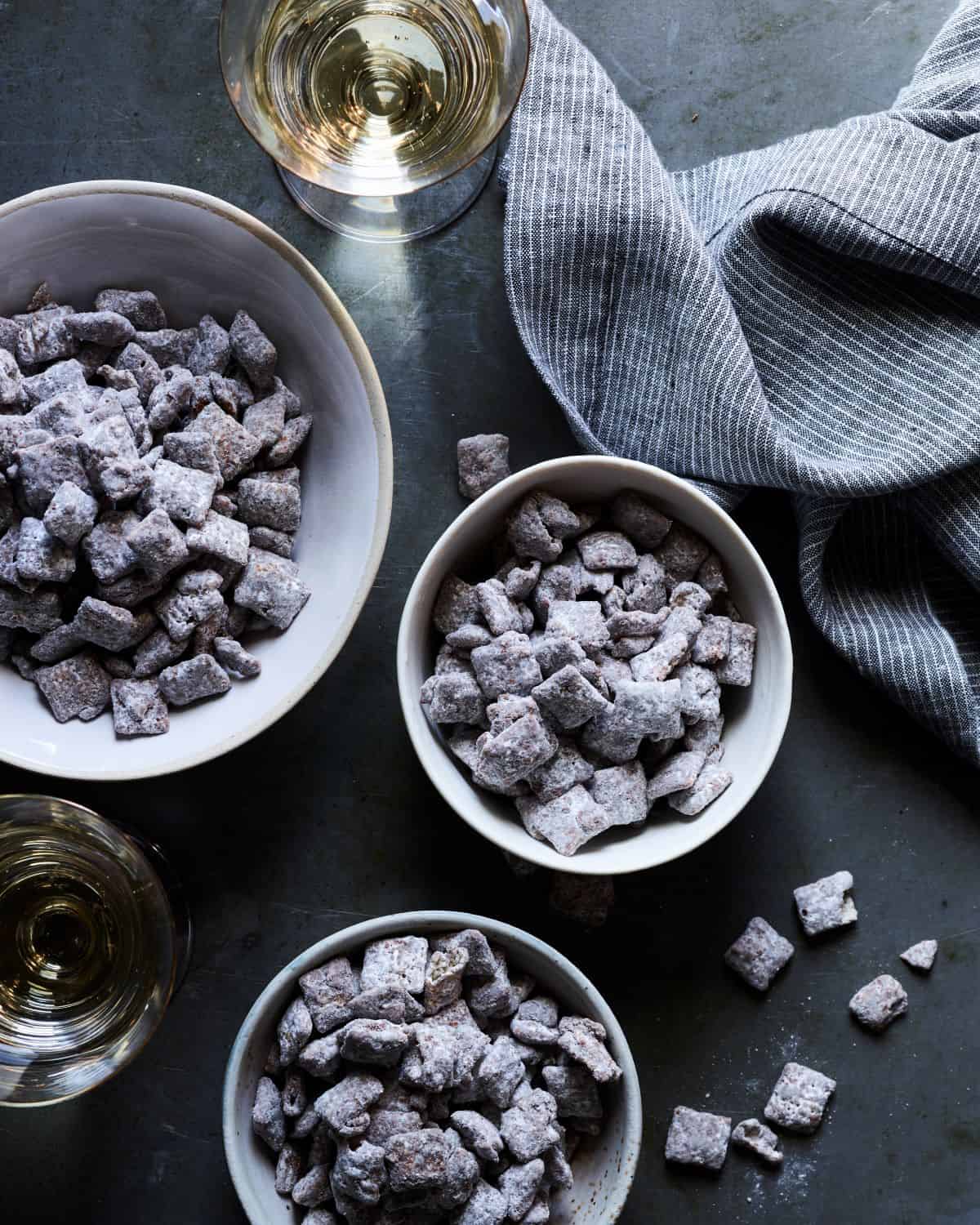 A big white ceramic bowl along with two smaller matching ceramic bowls filled with double dark chocolate muddy buddies, with two drinks and a striped grey-white kitchen towel.