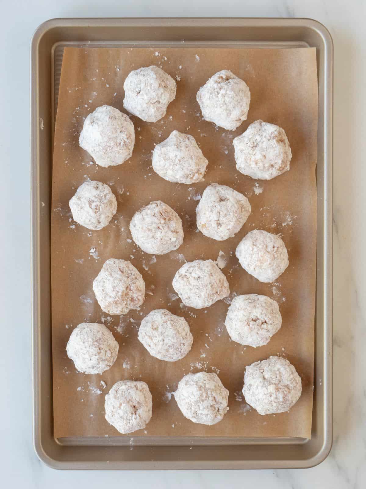 A parchment-lined baking sheet of chicken parm meatballs dredged in flour.