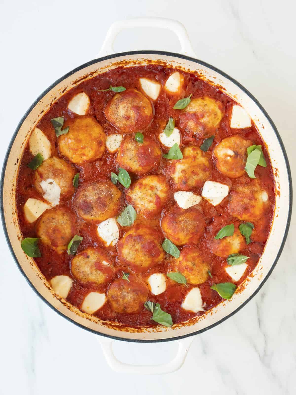A white dutch oven with chicken parm meatballs being cooked in tomato sauce garnished with pieces of bocconcini and basil leaves.