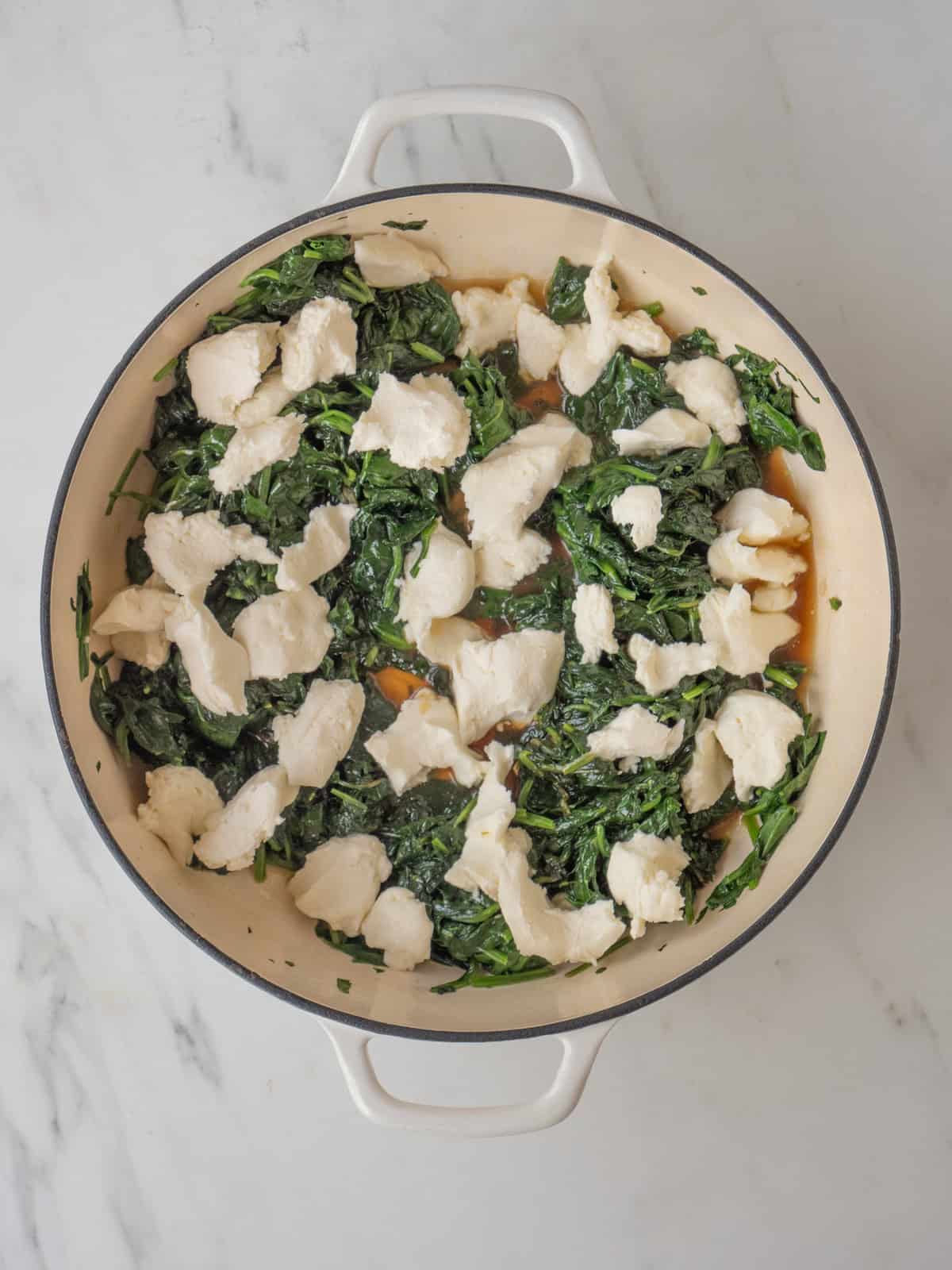 A dutch oven with cooked spinach, and cream cheese added in small pieces over it.