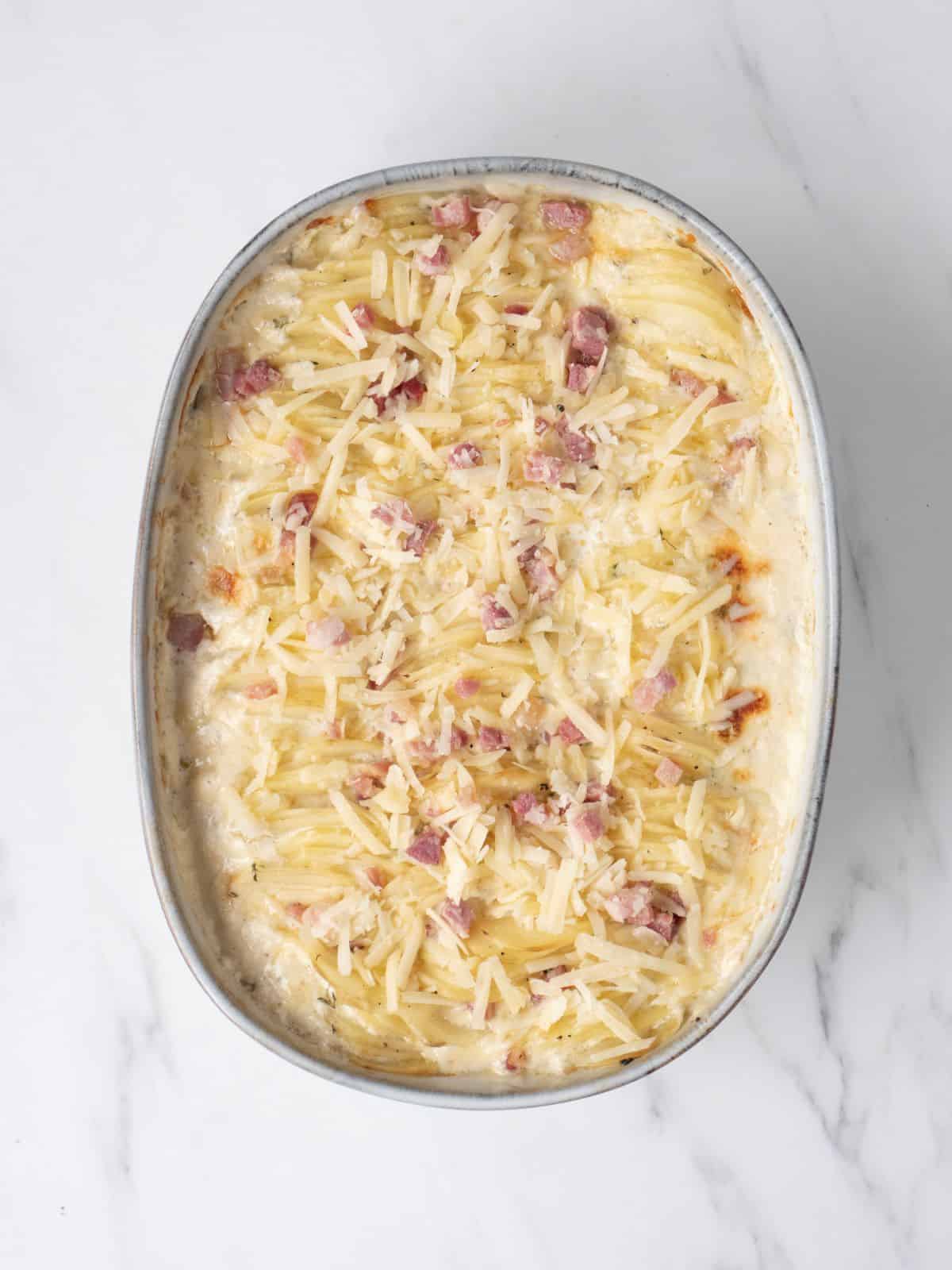 An oval baking dish with sliced potatoes dipped in cream mixture arranged in three columns along with the leftover cream mixture poured on top and the shredded cheese pancetta sprinkled on top.