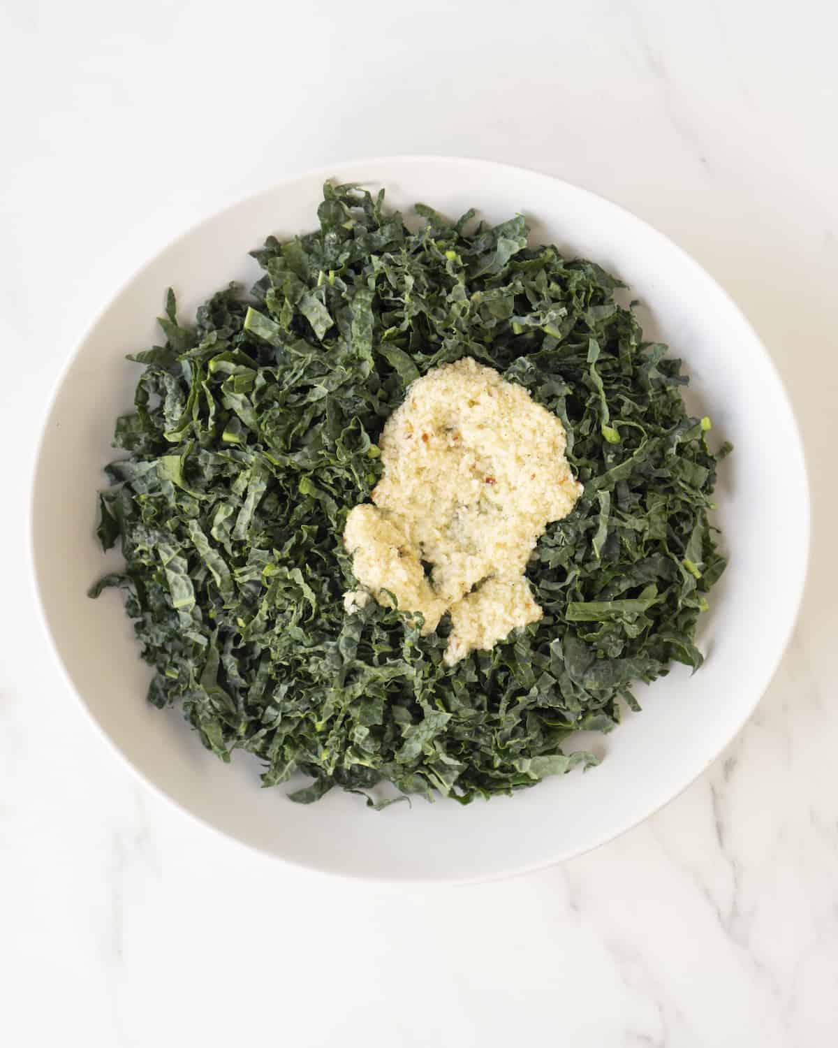 A ceramic white bowl of chopped kale and dressing on a white marbled countertop