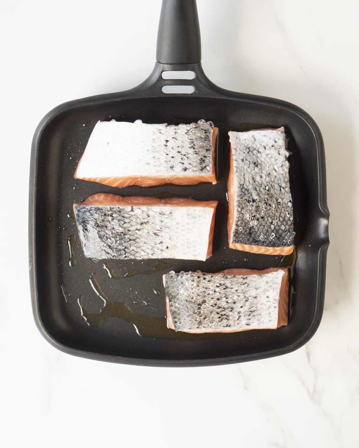 A pan layered with olive oil and salmon fillets cooking on a white marbled countertop. 