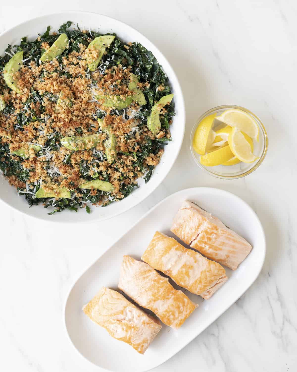 Kale salad in a white ceramic bowl beside a small clear bowl of sliced lemons and a serving platter of cooked salmon on a white marbled countertop.