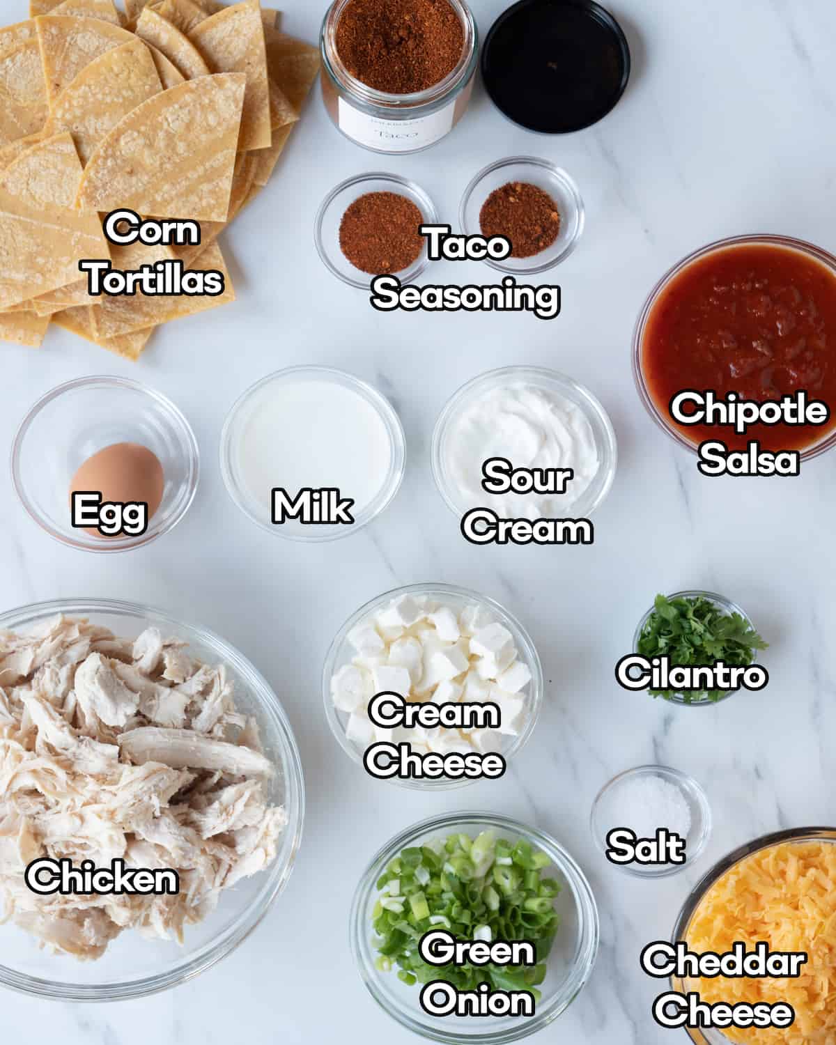 Ingredient shot of all ingredients in individual clear bowls including taco seasoning, corn tortillas, chipotle salsa, egg, milk, sour cream, cilantro, rotisserie chicken, green onion, cream cheese, and cheddar cheese on a white marbled countertop.