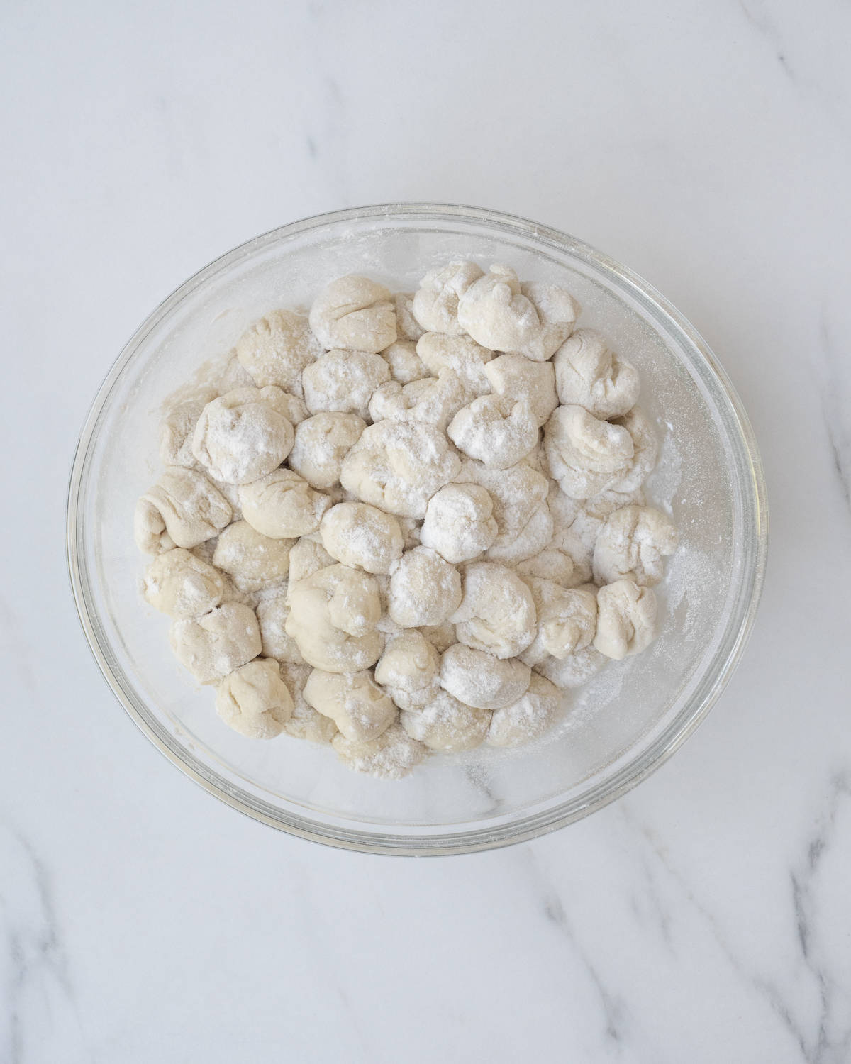 A clear white bowl of floured pizza dough separated in ½ inch cubes on a white countertop.