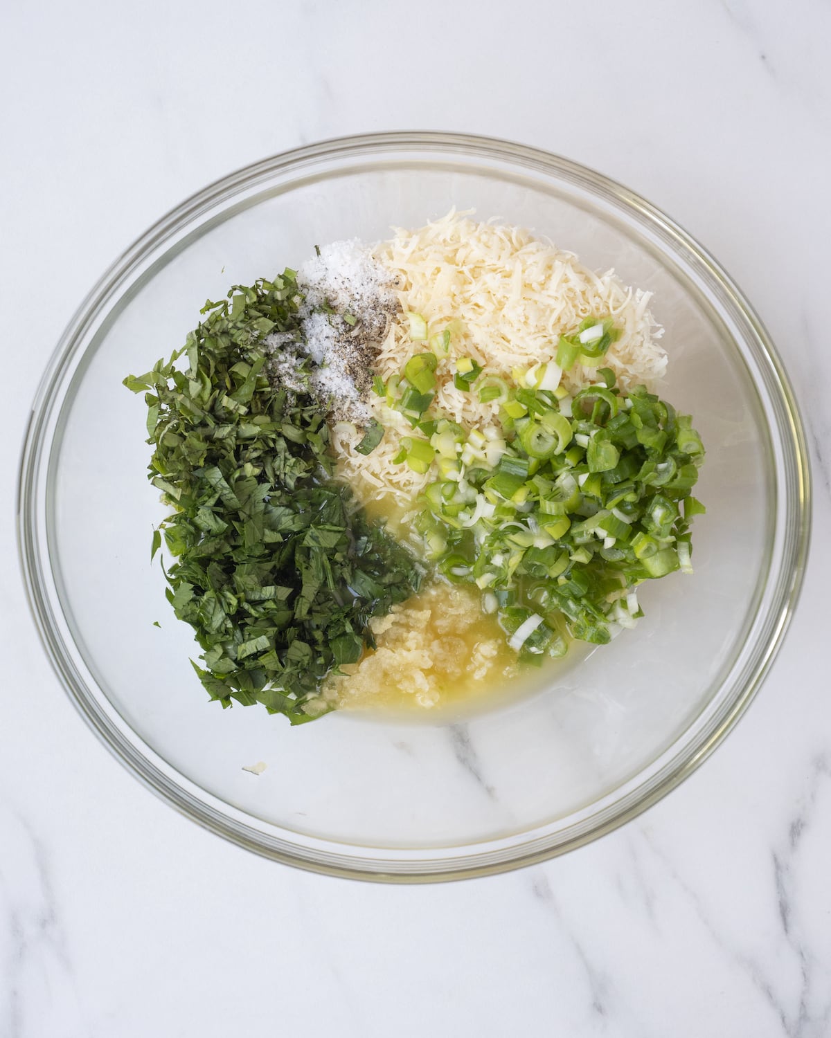 A clear white bowl of melted butter, scallions, parmesan cheese, parsley, garlic, and salt and pepper on a white countertop.