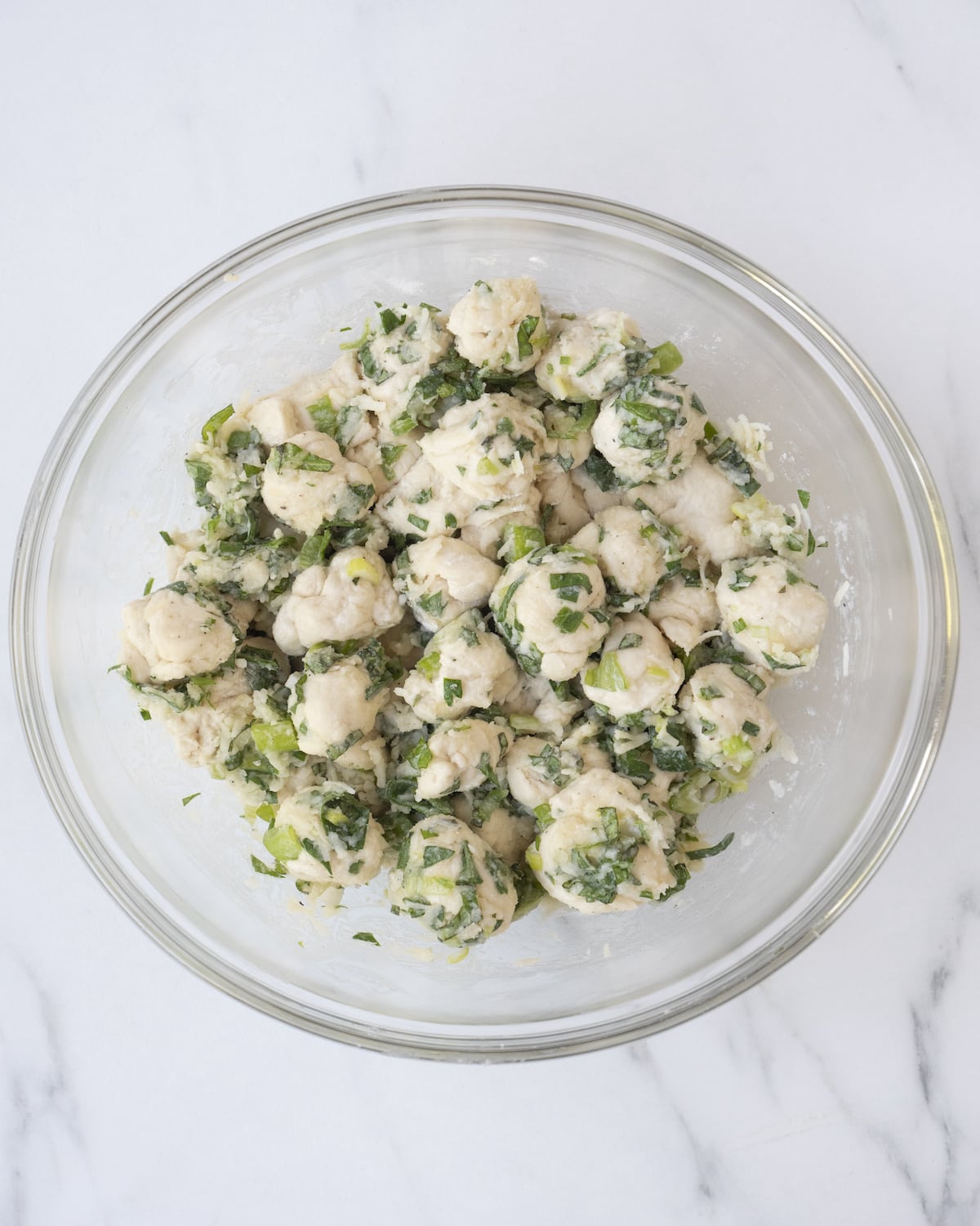 A clear white bowl of pizza dough mixed with herb butter mixture separated in ½ inch cubes on a white countertop.
