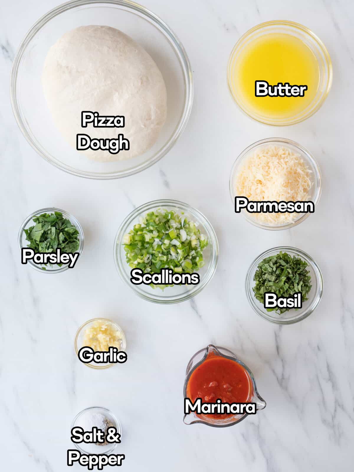 Ingredient shot of individual ingredients in clear bowls including pizza dough, butter, parmesan, basil, scallions, parsley, garlic, marinara, and salt and pepper on a white marbled countertop.
