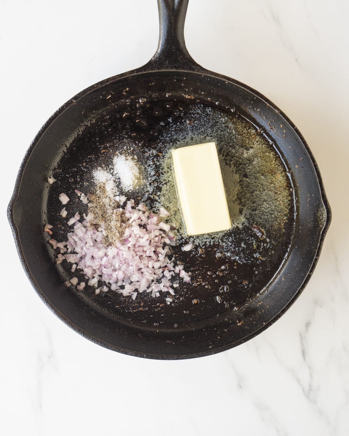 A cast iron skillet with butter, shallots, salt and pepper on a white marbled countertop.