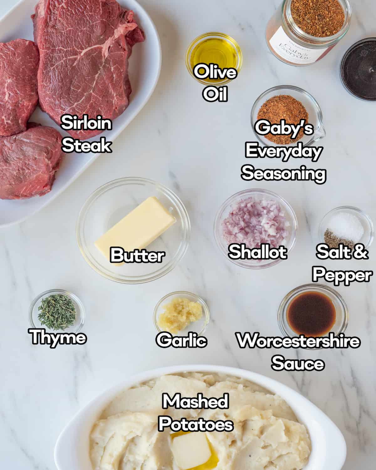 Ingredients in individual clear bowls including sirloin steak, Gaby's Everyday Seasoning from Dalkin&Co, olive oil, salt and pepper, shallot, Worcestershire sauce, garlic, butter, thyme, and mashed potatoes on a white marbled countertop.
