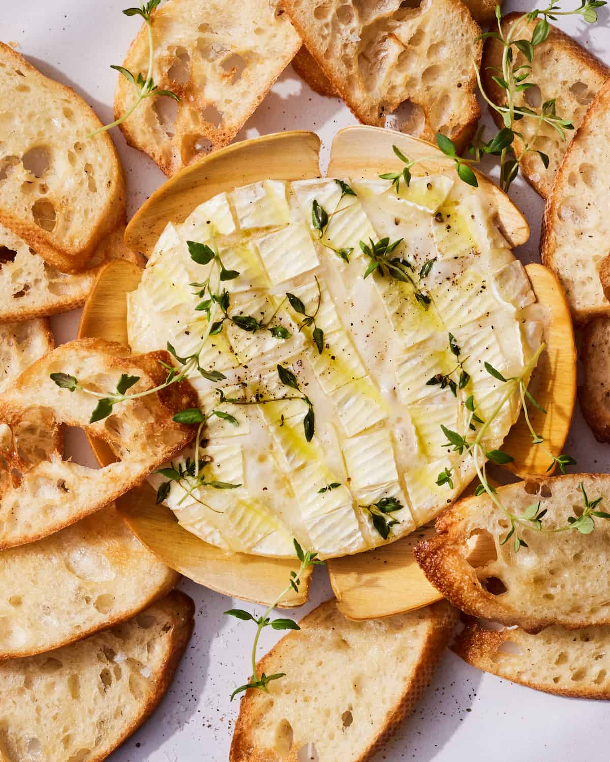 Thyme and garlic baked camembert on a wooden serving plate surrounded by bread slices topped with fresh herbs on a white countertop 