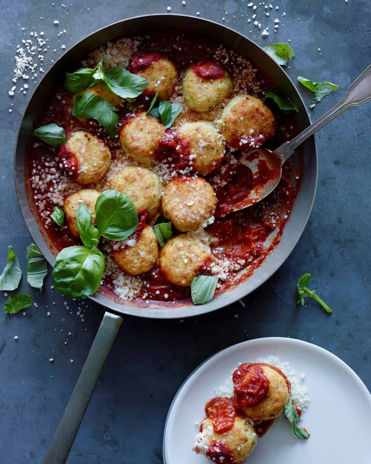 A skillet with chicken parm meatballs garnished with basil leaves and a serving spoon in the skillet, with a small plate with the meatballs in it on the side.