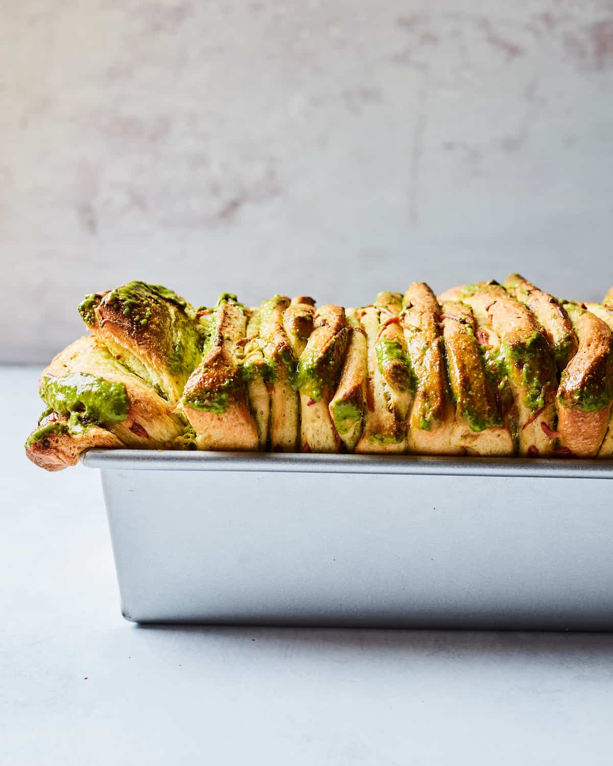 Pesto pull apart bread in a metal loaf pan on a white countertop with a white marbled background