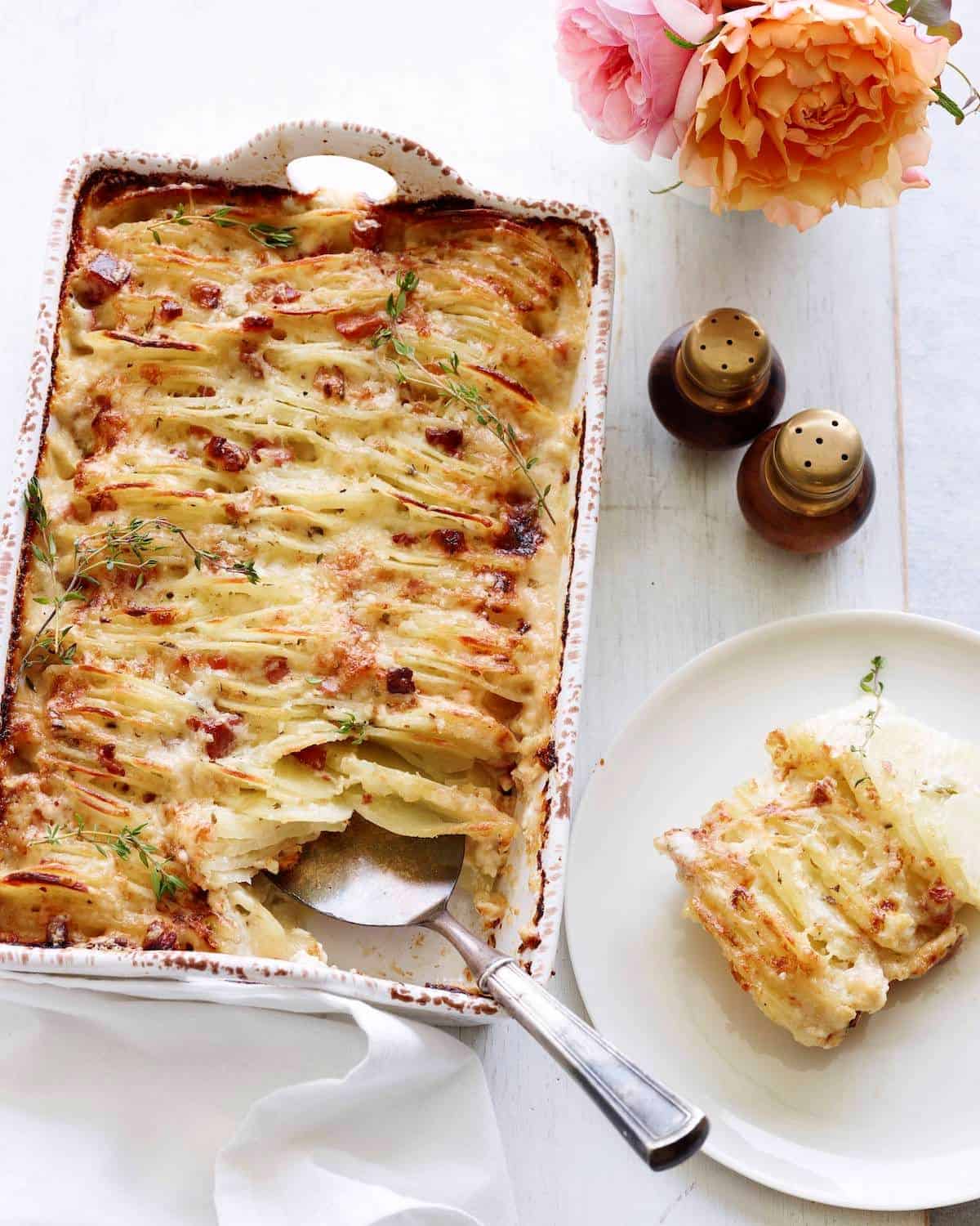 A rectangle white baking dish with hasselback scalloped potatoes baked dish with a flat spoon to serve, and a corner piece served into a white plate on the side, along with some flowers in a vase and salt and pepper shakers on the side.