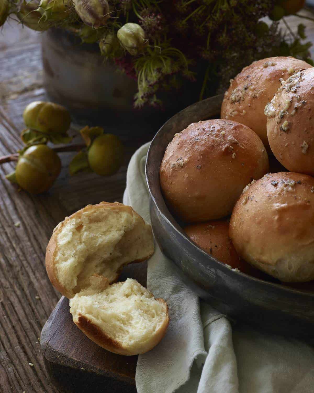 Homemade garlic herb rolls in a metal serving dish on top of a light green linen napkin and wooden cutting board with floral foliage behind the dinner rolls on a wooden countertop 