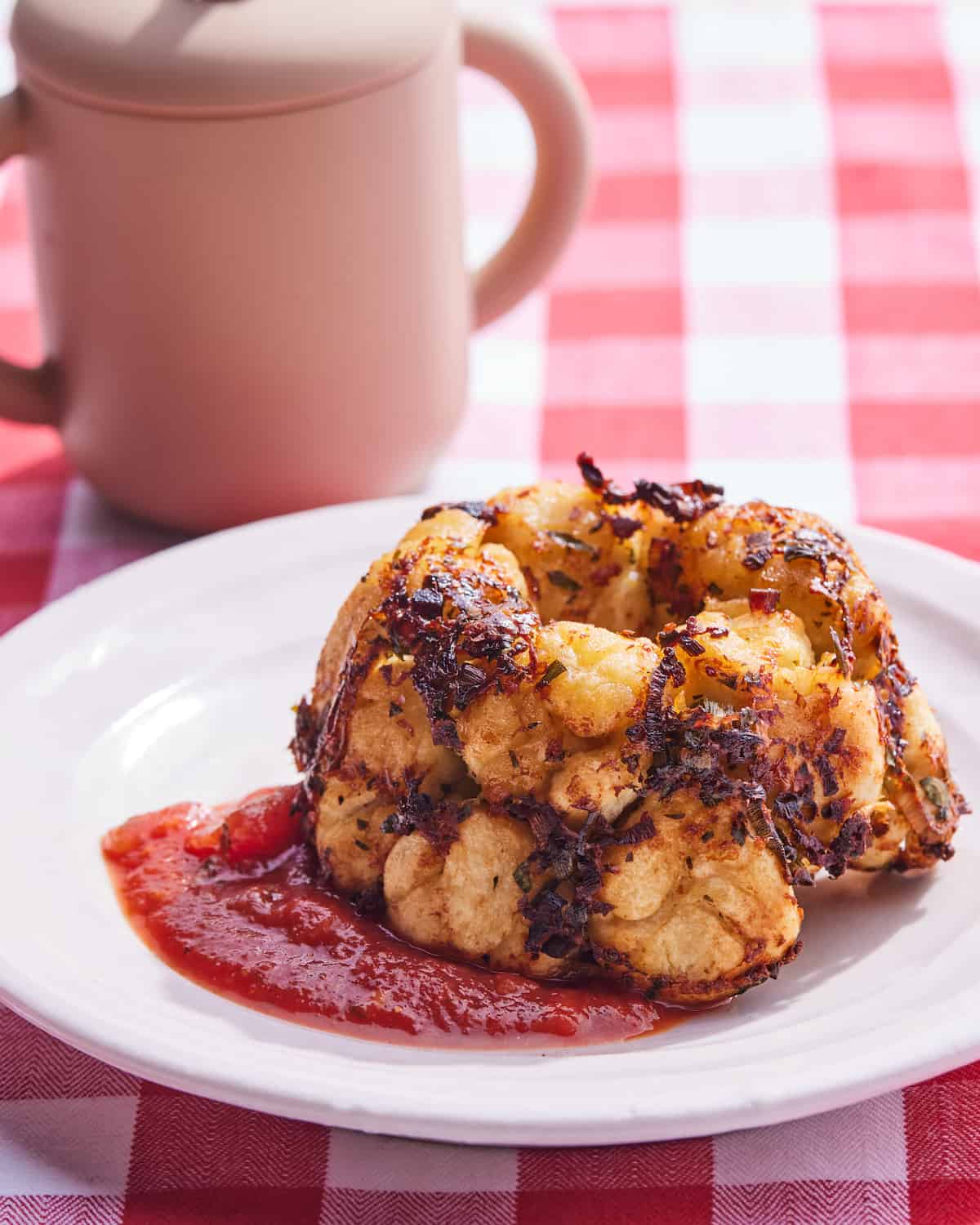 A piece of Pizza Monkey Bread and marinara sauce on a white ceramic plate in front of a pink sippy cup on a red and white checkered table cloth.