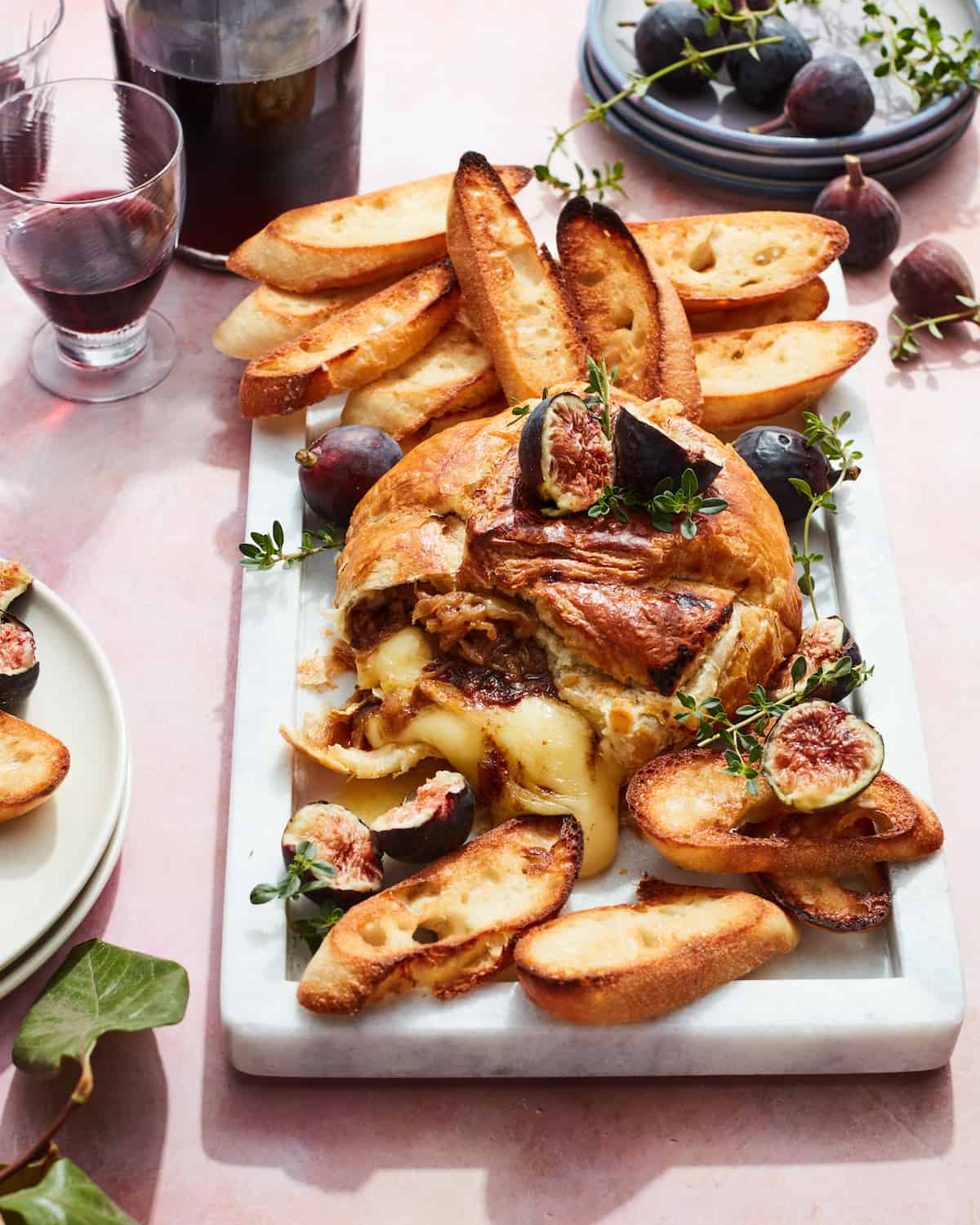 A  white marble rectangular platter with puff pastry wrapped baked brie, melting out of the puff pastry, baguette slices, thyme sprigs, halved figs along with some stacked plates, wine, a wine glass and more whole figs on the side.